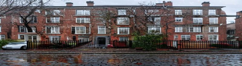 Panoramic view of a terrace of red-brick Dublin Georgian houses with sash windows and brightly painted front doors, cast-iron railings, wet cobblestone street reflecting grey morning light, a single parked estate car, bare tree in the foreground, soft Irish spring drizzle, no people no faces no hands
