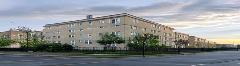 Panoramic exterior view of an Irish institutional accommodation building at dawn, multi-storey beige render with rows of small windows, asphalt yard with parking spaces, low chain-link perimeter fence, leafy hedgerow boundary, soft Atlantic morning light, low cloud cover, no people no faces no hands