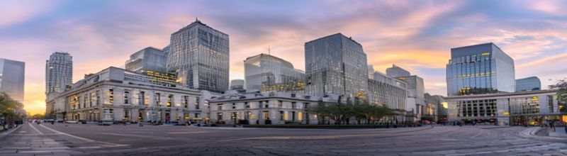 Panoramic view of a modern London financial district skyline at early morning, Bank of England&rsquo;s classical stone facade in the foreground, glass towers of the City rising behind in soft haze, lights still on in upper office floors, empty cobbled street, pale yellow sunrise light, no people no faces no hands