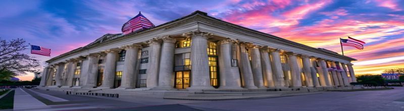 Panoramic wide-angle photograph of a U.S. Treasury Department building facade at dawn, neoclassical limestone columns, brass doors, American flag on a tall flagpole catching first light, documentary realism, no people no faces no hands