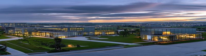 Panoramic view of a modern grey-glass corporate campus on the outskirts of an Irish city at dusk with a wide landscaped lawn, three low-rise office blocks lit warm from inside, a polished granite IDA Ireland-style monolith sign at the entrance, asphalt access road curving into the foreground, distant Wicklow-style hills in pale silhouette, no people no faces no hands