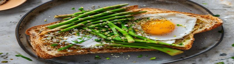 Panoramic overhead view of a thick slice of toasted sourdough bread on a slate kitchen plate, a bundle of bright green asparagus spears laid across it, a glossy poached hen egg with the yolk just broken, a scattering of golden breadcrumbs and chopped wild garlic on top, a small dish of brown butter to the side, soft warm late-afternoon kitchen light, sharp natural detail on the toast crust and asparagus tips, no people no faces no hands